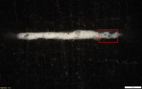 Black and white photograph. A detailed view of a rock or mineral surface which has a textural black appearance, and a strand of white running horizontally across it. On the lower left yellow text reads, ‘Magnification: 12.6 x’. On the right, a small white rectangle bears the number 100.