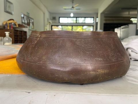 Colour photograph. Documentation of a low, wide vessel or container resting on a table top, seen straight on. The vessel is a warm, bronze brown colour, with a series of flowing, organic patterns scratched into its surface, and a slightly worn patina.