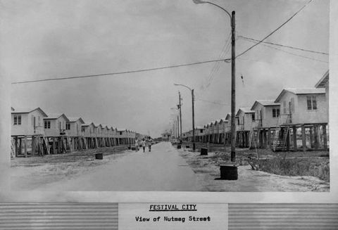 Black and white photograph. A view down a wide street, lined on either side with rows of beach huts on raised stilts, with steps leading up the entrance. Tall street lamps line one side of the street. On the right, three people can be seen in the distance, walking towards the camera. Below the photograph, a cut-out passage of text in the lower centre reads, ‘Festival City, View of Nutmeg Street’.