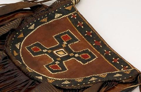 Colour photograph. A close-up documentation of a brown and black, semi-circular leather bag or garment adorned with ornate and intricate patterns. A series of colourful geometric patterns have been woven onto the surface of the bag, including an angular, central motif with elements of black, red and white. Leather fringing can just be seen around the curved edge of the bag.