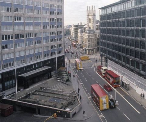 Colour photograph. A view from an upper window looking down onto a busy city street below, filled with cars, buses and people. On the left and right of the foreground we see two modern, multi-storey buildings, while a cathedral is visible in the distance.