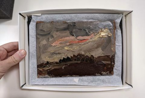 Photograph of painting shown resting on foam and tissue paper in a cardboard archival box. On the left, a person's hand holds the box, their thumb measuring almost half the height of the painting.