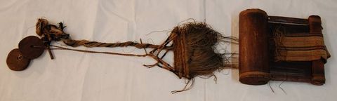 Colour photograph. Documentation of a wooden hand loom laid out across a white background. The loom is made from elements of dark wood and brown and black twine, and contains a small panel of brown, black and red woven fabric. 