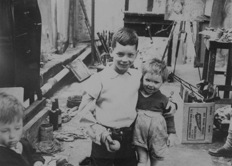 Black and white photograph. Two young children in an artist’s studio smile at the camera. The elder has a protective arm around the younger, and holds a ball in his other hand. On the far left, the head of a third child is just visible, and on the right, we see the cropped foot of another person. Behind them is evidence of an artist’s studio including paint brushes, paint cans, and two easels.