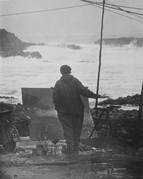 Black and white photograph. An artist is seen from behind looking out to the stormy sea beyond, facing an artwork leaning on an easel. They hold one tool out in their right hand, above a tripod table. On the ground, art materials have been laid out in front of the artwork. On the left, we can just make out the rear end of a motorbike.