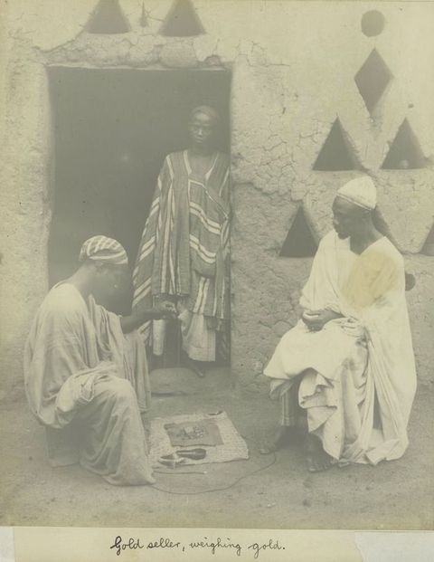 Black and white photograph. Three people are positioned near an open doorway; one leans against the doorway, while the other two are seated on small stools. They are all dressed in loose, draping fabric. Between the two seated people is a rectangle of fabric containing a series of objects. Below the photograph, a panel of handwritten text reads, ‘Gold seller, weighing gold.’