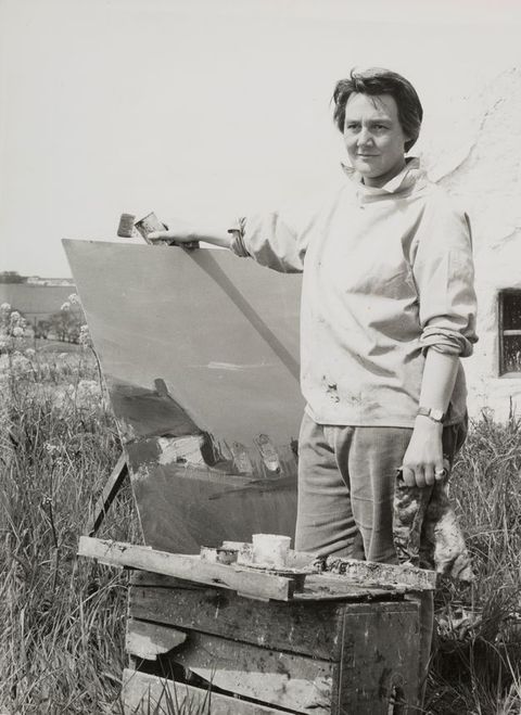 Black and white photograph. A woman dressed in loose, paint splattered clothing stands in an outdoors space facing towards the camera, with eyes averted into the distance. She grips an artwork resting on an easel with one hand, and in the other she holds a rag. In front of her is a wooden box on which art materials rest. Behind her, the wall of a building is just visible, along with long dry grasses rising up from the ground.