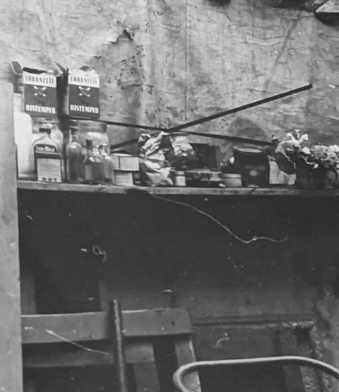 Black and white photograph. The partial view into a warehouse or studio space, showing a busy shelf crowded with bottles, jars, and containers. Behind the shelf we can see a ripped piece of suspended fabric. Below the shelf, various pieces of furniture have been leaned against the wall.