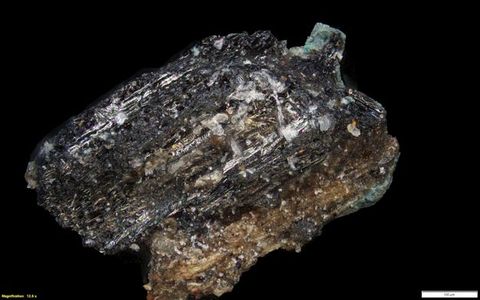 Colour photograph. A close-up, detailed photograph of rock, crystal, or mineral with a roughly textured, glistening surface, against a black background. On the lower left yellow text reads, ‘Magnification: 12.6 x’. On the right, a small white rectangle bears the number 100.