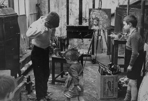 Black and white photograph. Three young children stand in an artist’s studio, with heads turned away from the camera. They are surrounded by messy detritus including paint brushes, paint cans, and a wooden box filled with junk. On the right, two easels can be seen, one with a small painting of a child resting on it. The cropped head of another child is just visible on the far left. 
