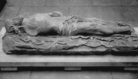 Black and white photograph. Stone statue of a slim, reclining man spread across draped fabric, seen from an elevated viewpoint. A small loincloth covers his groin area. The man has long hair and a beard, with eyes closed and lips slightly parted. His arms and feet have broken away.