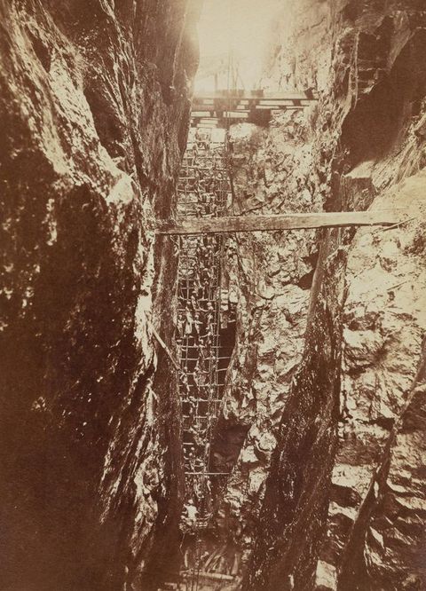 Sepia photograph. A sepia-tinted photograph in portrait format, showing the view into a steep mine shaft from side on, with sharp, rocky surfaces flanking a steep recess. Metal mesh covers part of the rocks, and wooden planks run horizontally across the image.