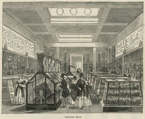 Drawing. This artwork shows the view into a high-ceilinged museum space filled with glass display cases containing Egyptian mummies and other treasures, and Egyptian artefacts hung onto the walls. People dressed in traditional clothing including top hats, suits, bonnets, and long skirts, walk between the exhibits. Below the illustration reads the text, Egyptian Room.’