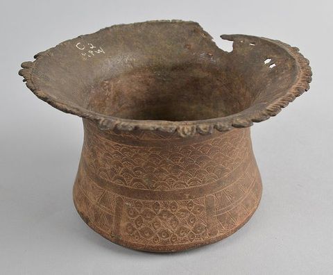 Colour photograph. Documentation of a pot or vessel seen from above with a wide, fluted outer brim that is partially worn away. The outer surface has a series of patterns scratched into its surface, and the outer brim has a delicately decorated trim. The rim is inscribed with white letters or numbers.