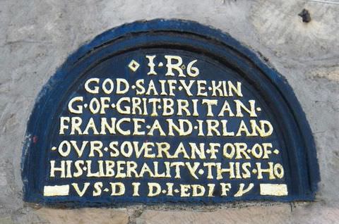 Colour photograph. Documentation of a blue wall plaque in a semicircular shape, decorated with a passage of white, upper-case text in old English. The plaque sits on a grey background. Text includes the passage, ‘GOD SAIF YE KIN’. 