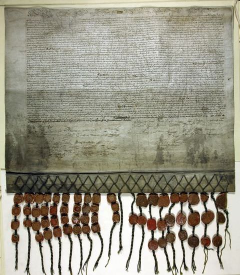 Colour photograph. A document of a fabric wall hanging adorned with a long passage of cursive text. Black stitches run along the lower edge of the fabric, with dangling pieces featuring rows of brown beads.