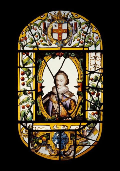 Colour photograph. A photograph of an oval, backlit stained-glass window featuring a colourful, intricate design adorned with plants, berries, and a crown. Within an oval in the centre of the design is a head to shoulder portrait of a man wearing sumptuous, shiny fabric and a stiff white collar. 