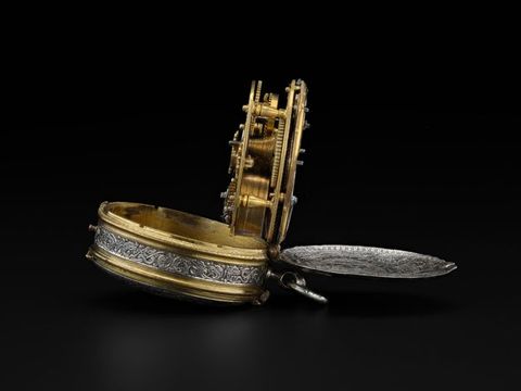 Colour photograph. Documentation of a gold and silver object with mechanical, clock-like details, seen from a side-on view against a black background. The lid and inner core of the object have been pulled open. 