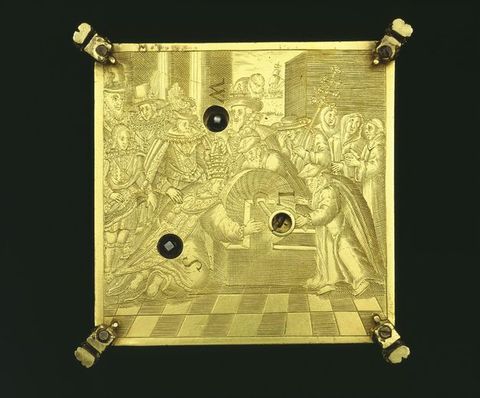 Colour photograph. A document of a gold, square object against a black background. An illustration embossed into the object shows a crowd of people in an interior space gathered around a cylindrical object, on a chequered floor. Each corner of the object has a clamp-like fixture, and within the square are three small, circular holes.