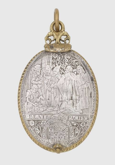 Colour photograph. A silver, oval object featuring intricate, highly detailed decoration, photographed against a white background, with gold details running around its trim. The silver face is adorned with an illustration featuring a crowded cluster of people, and the royal British insignia below.