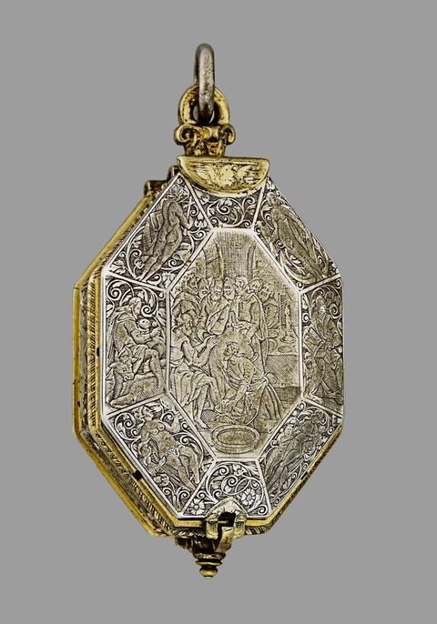 Colour photograph. An octagonal, three-dimensional object is photographed against a pale grey background. The object has a silver surface and gold detailing around the edges. Its surface is embossed with a series of decorative patterns. 