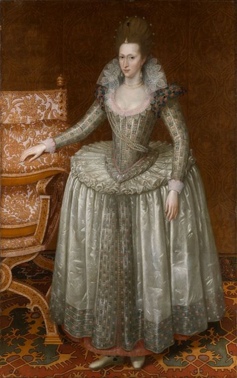 Painting. A full-length portrait showing a formally dressed woman in an indulgently-decorated interior. The room is decorated in shades of brown and beige. She wears a white, an embroidered dress with a long, full skirt, a tight corset, and an upright white lace collar. Her right hand rests on an intricately patterned chair to the left of the image.
