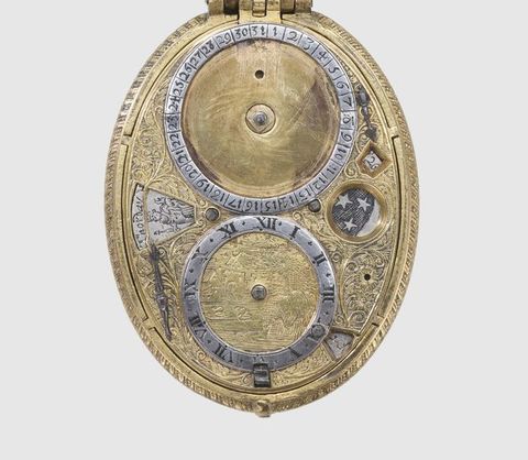 Colour photograph. The inside, lower lid of an oval object is photographed against a white background. The hinge connecting it with the upper lid is just visible at the top. The object features cylindrical and numerical details resembling clock or compass parts. 