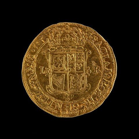 Colour photograph. A shiny gold coin photographed against a black background. The coin is inscribed with text running around its circumference, and the royal British insignia, featuring a lion and a harp, in the centre.