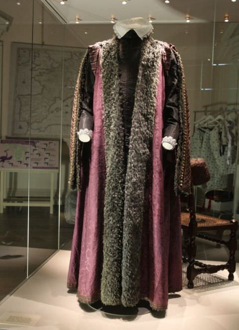 Colour photograph. A historical costume comprised of a long, purple cape-like, sleeveless coat with a brown fur trim, worn over a brown, fitted underlayer, is displayed on a headless and handless mannequin, which sits within a larger, glass display box. In the room beyond, further display items include maps and clothing on a rail.
