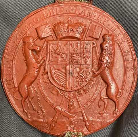 Colour photograph. A burst red circular object photographed against a grey fabric background. The object features a low-relief carving of the royal British insignia, with the lion, the unicorn, and the crown, clearly visible. 