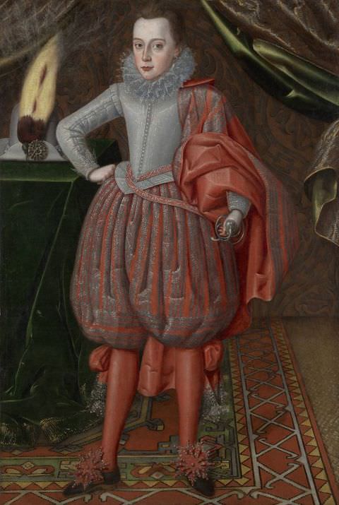 Photograph. A young man dressed in historical clothing including a corset, pantaloons, a stiff collar, and red tights, stands within an interior featuring indulgent fabrics and a detailed rug. In the distance, curtains can just be seen sweeping outwards on the left and right, while on the left, a tall, white hat decorated with a feather sits on a table adorned with a green velvet blanket.
