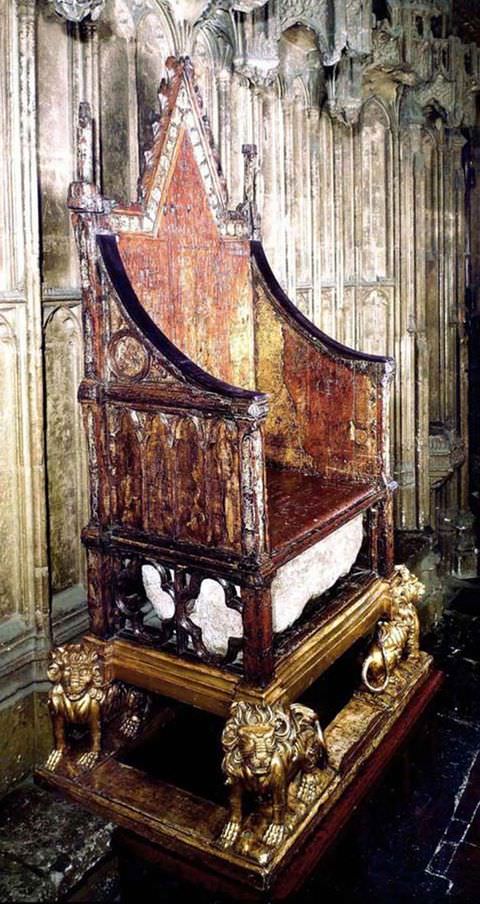 Colour photograph. Documentation of a tall, brown wooden throne seen from an oblique, side-on view. The throne sits on four feet in the shape of standing lions and has a tall, pointed back, while its sides form curved slopes. 