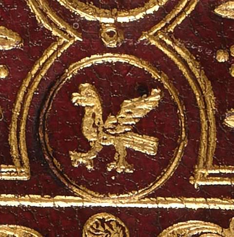 Colour photograph. A close-up photograph showing a detailed, gold decoration embossed into a red ground. The centre of the decoration features a circular shape containing a stylised bird with large head and feet.