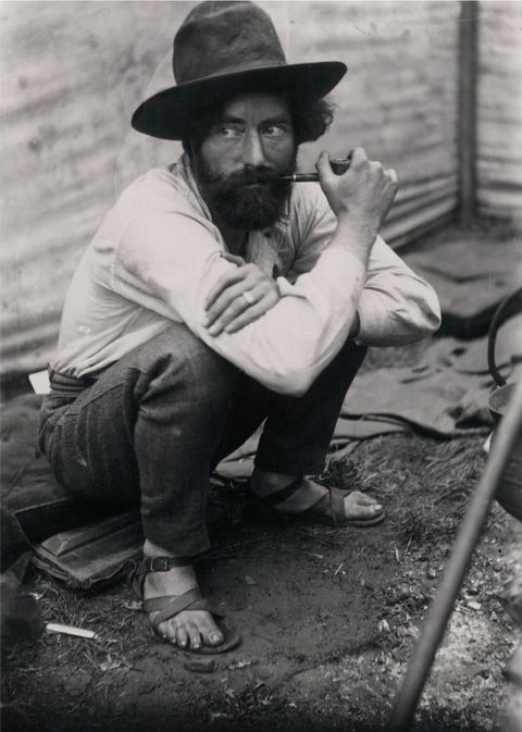 Black and white photograph. A man dressed in a white shirt, jeans, sandals, and a large hat holds a pipe in one hand and squats on a mattress just visible on the dusty outdoor ground.
