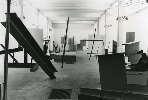 Black and white photograph. The view into an interior space filled with metal sculptures that seem to lean in contrasting directions. The partial silhouette of a man can be seen on the left of the image, and on the right a man is seated in the shadows behind a desk.