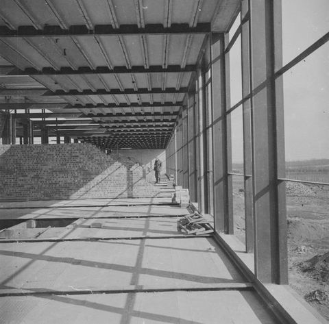 Black and white photograph. The perspectival view into an interior space which appears mid-construction, with partially covered floors, exposed ceiling beams and partial brick walls. A standing person can be seen in the far distance.