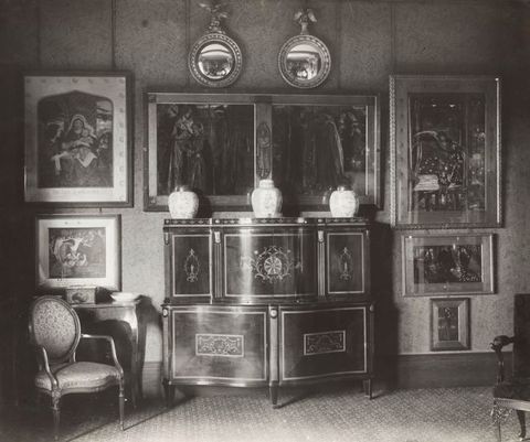 Black and white photograph. The view into an interior featuring old, ornate furniture. A central cabinet has three urns resting on it, and is surrounded by works of art in rectangular and circular frames. Two chairs are visible on the left and right.