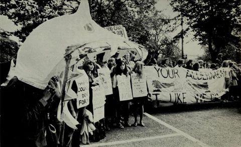 Black and white photograph. A crowd of people stand side by side holding fabric and paper signs bearing text slogans. To the left, a group of people are covered by a curved canopy shaped like a shark, with jagged teeth. Trees can be seen in the distance.