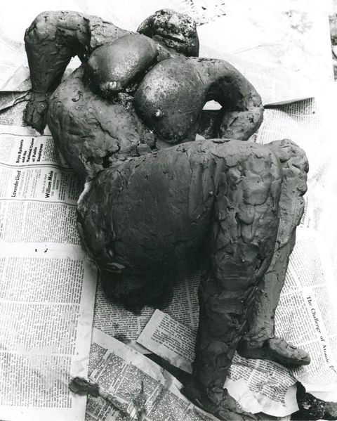 Black and white photograph. An abstracted, expressive sculpture of a nude figure seen from above, lying across a floor strewn with newspaper.