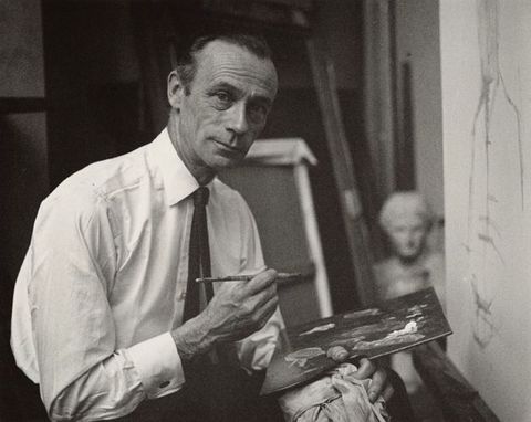 Black and white photograph. A photographic portrait of a man from waist up in a white shirt and tie, who looks to the camera with a slight smile. He balances a paint palette in one hand, and holds a paintbrush in the other.