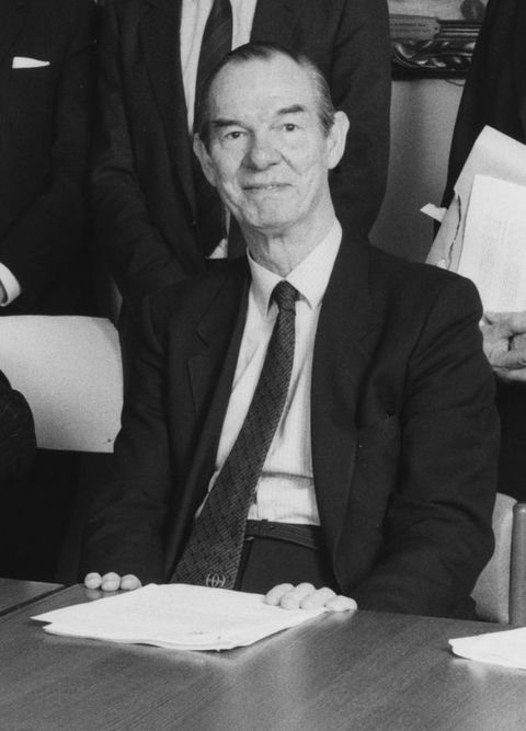 Black and white photograph. A man wearing a suit is seen from the waist up, with a desk before him bearing paper. Chairs can just be seen around him, and the torso of a standing man in a suit is visible behind him.