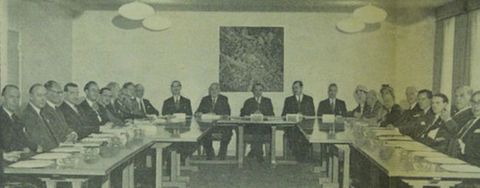 Black and white photograph. A group of smartly-dressed men face towards us, seated around a u-shaped table. A square, abstract artwork hangs on the wall behind them, while bulbous lamps are suspended from the ceiling.