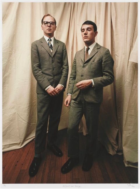 Colour photograph. Two smartly dressed men in stiff grey suits stand on natural wood floarboards, and a white curtain can be seen behind them. The taller man, seen on the left, wears glasses.