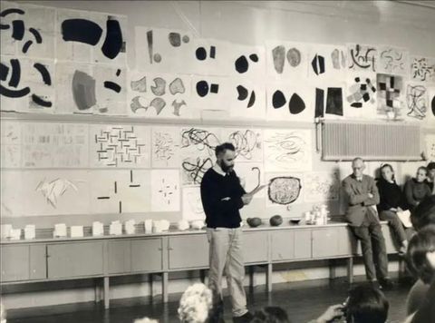 Black and white photograph. A man in a dark sweater and pale trousers looks down at paper in his hand, while standing in front of a wall emblazoned with abstract works of art. People can just be seen around him listening to him speak.