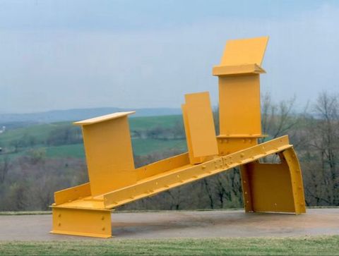 Colour photograph. A vivid yellow sculpture made from geometric panels of metal is seen in an outdoor setting, with softly focussed grass, trees and hills visible in the distance.