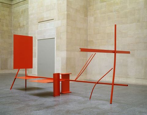 Colour photograph. A bright orange sculpture made from geometric panels of metal sits within an interior space, featuring a grey floor and stone walls.