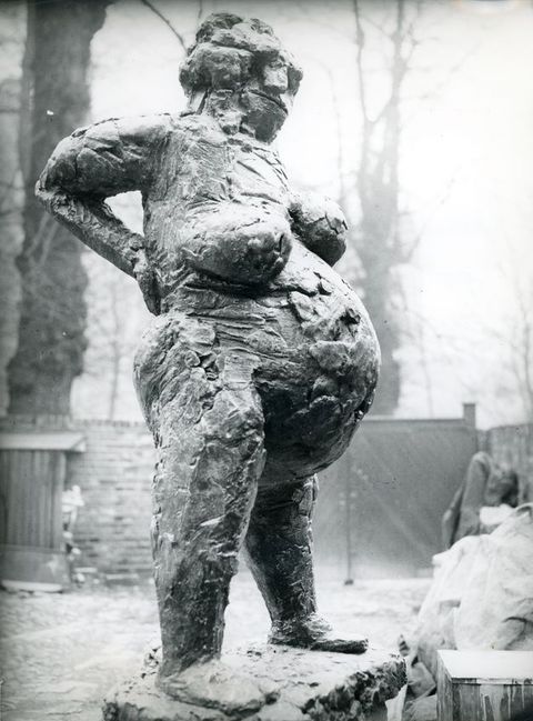 Black and white photograph. A sculpture of a nude, pregnant woman with a rough, expressive surface is seen in an outdoor setting, with trees, walls and a gate just visible in the distance.