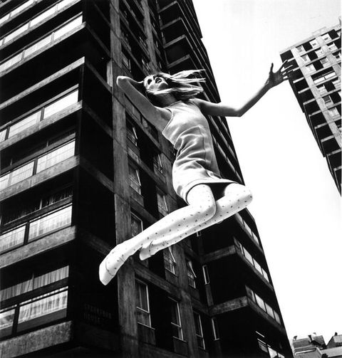 Black and white photograph. A young woman wearing a mini dress and polka dot tights is seen from below, and appears suspended mid-air, while perspectival buildings can be seen behind her.
